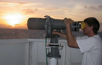 An observer uses high-powered binoculars to scan for marine mammals from the deck of a ship.